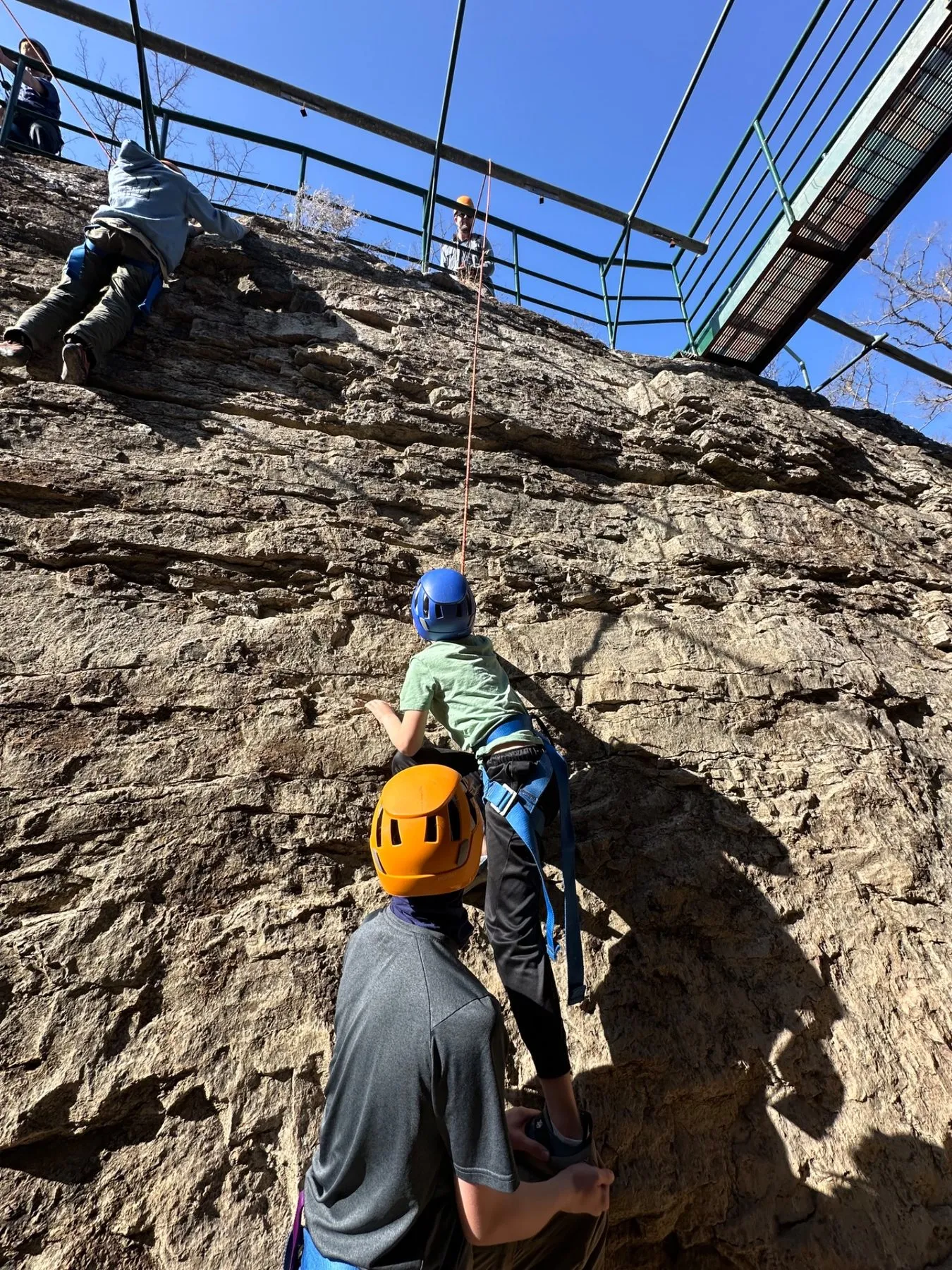A Scout climbing a rock face with a belay team below
